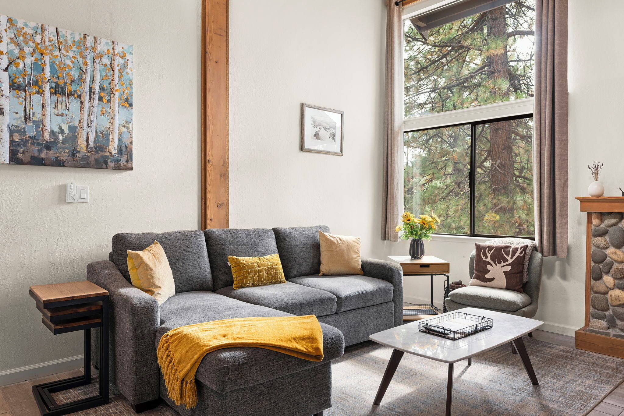 A living room with a gray sofa, a coffee table, a painting, and a window with a view of trees.