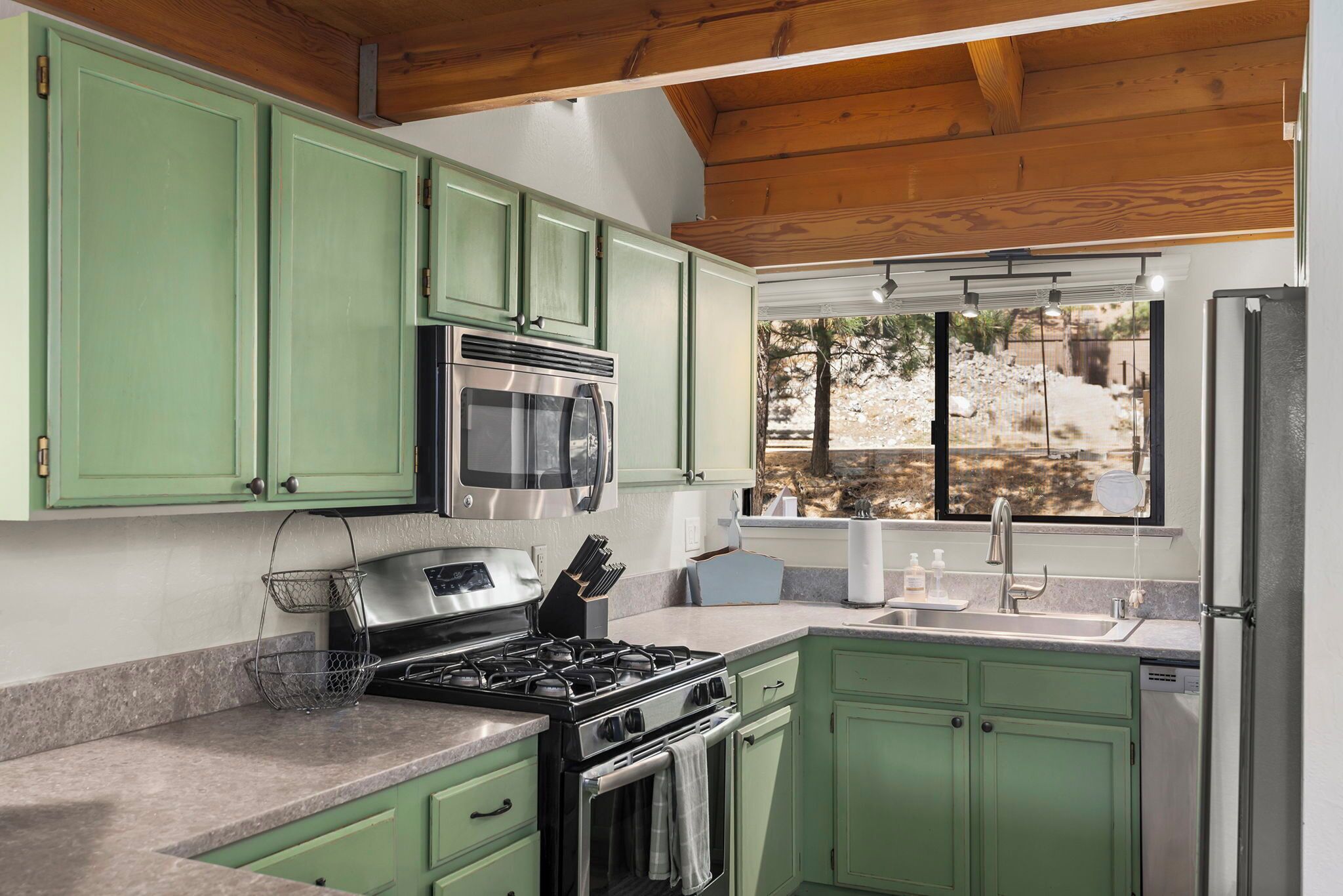 Cocina con gabinetes verdes, electrodomésticos de acero inoxidable y una ventana con vistas a un paisaje rocoso.