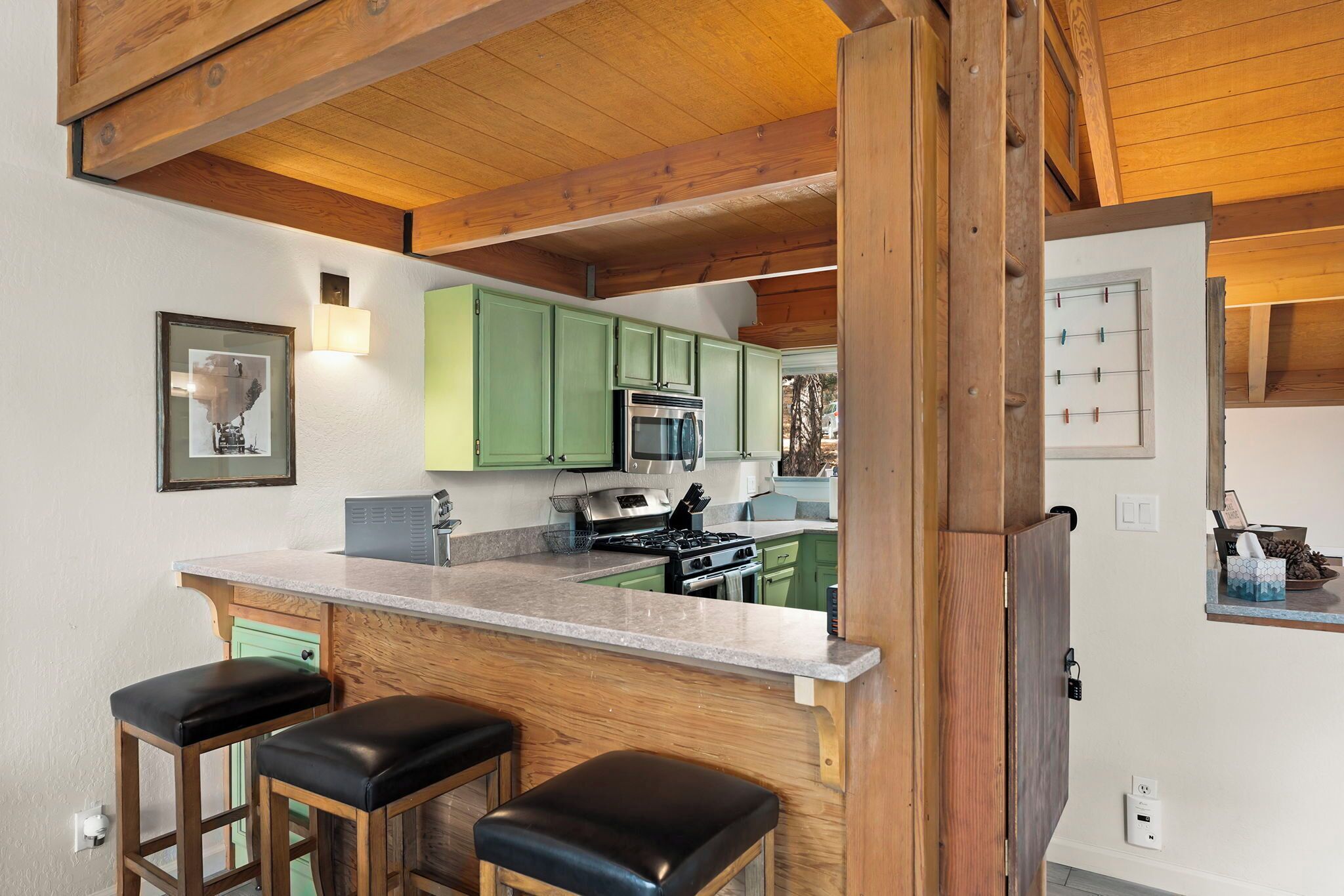 Une cuisine avec des meubles de couleur verte, une cuisinière en acier inoxydable et un plafond en bois. On y trouve deux tabourets de bar avec des assises noires.