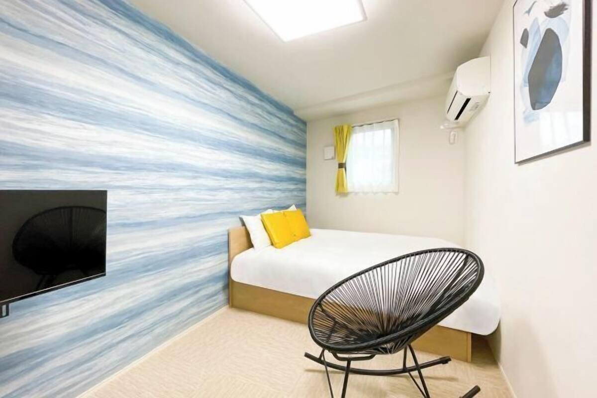 A bedroom with a bed, a black chair, a flat-screen TV, and a wall with a blue and white abstract design.