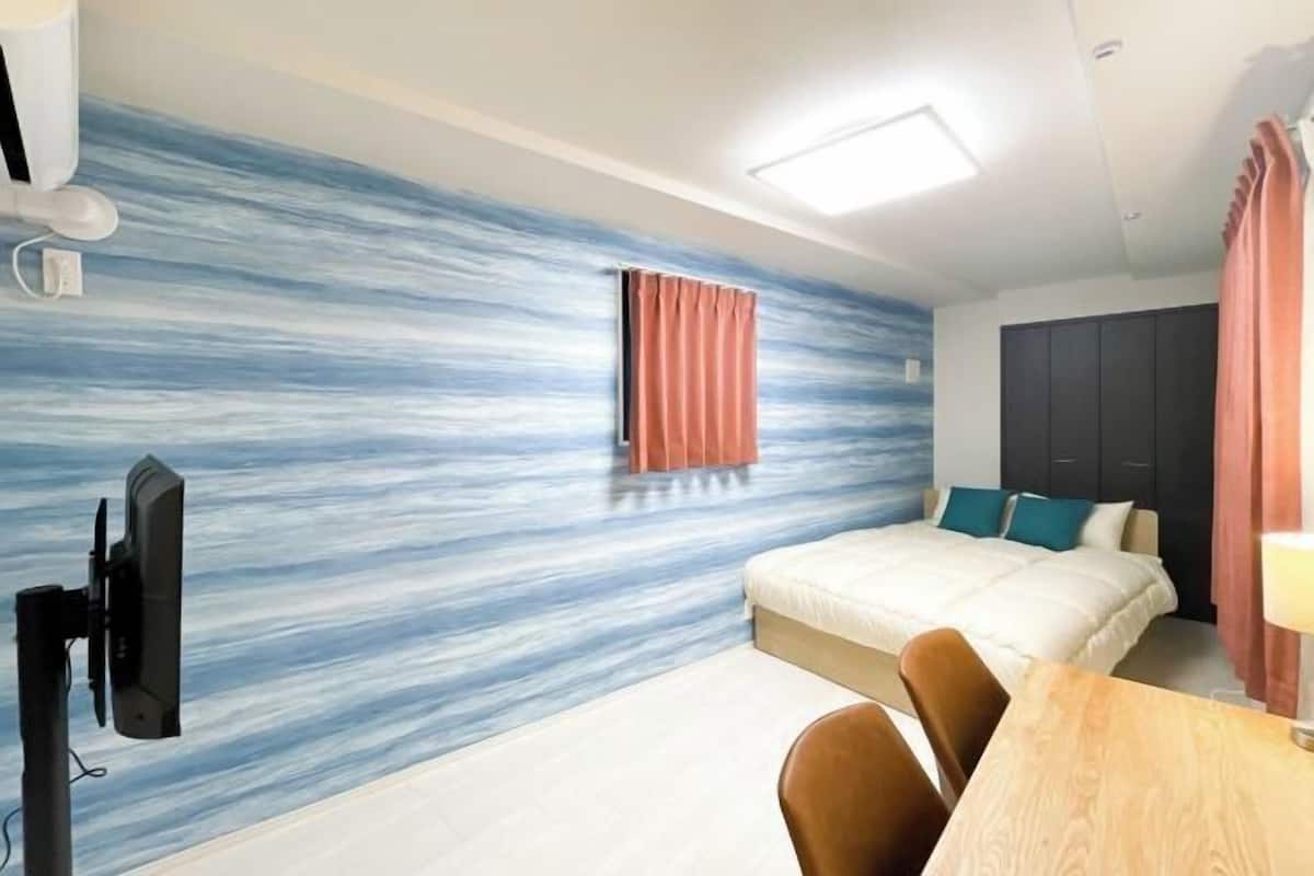 A hotel room with a bed, a desk, a chair, a TV, and a wall with a blue and white pattern.