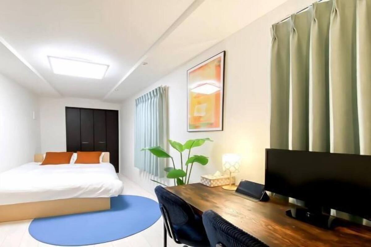 A modern hotel room with a bed, a desk with a flat-screen TV, a blue rug, and a plant.