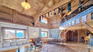 A spacious wooden interior with a high ceiling, a dining area, and a sofa set by a window with a mountain view.