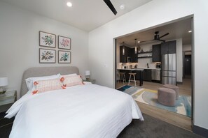 A modern bedroom with a large bed, a small kitchenette, and a bathroom visible through an open door.