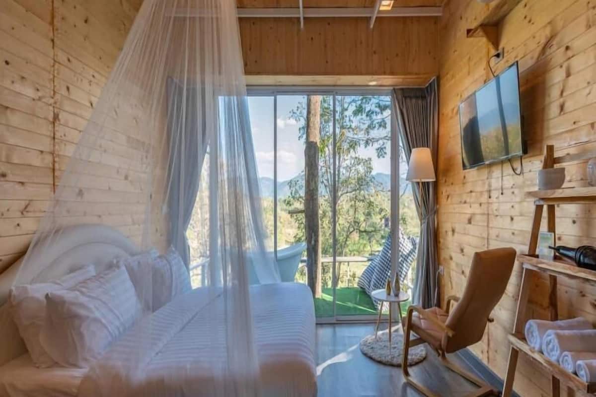 A wooden cabin room with a bed, a chair, a TV, and a view of the outdoors.
