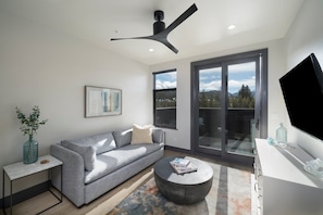 Un salon moderne avec un canapé gris, une table basse ronde, un téléviseur posé sur un meuble blanc et un ventilateur de plafond.