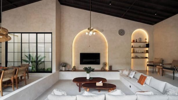 A spacious living area with a large sofa, a coffee table, and a TV mounted on the wall. There are shelves with decorative items and a dining area with chairs and a table. - MDF Villa Canggu (Bali)