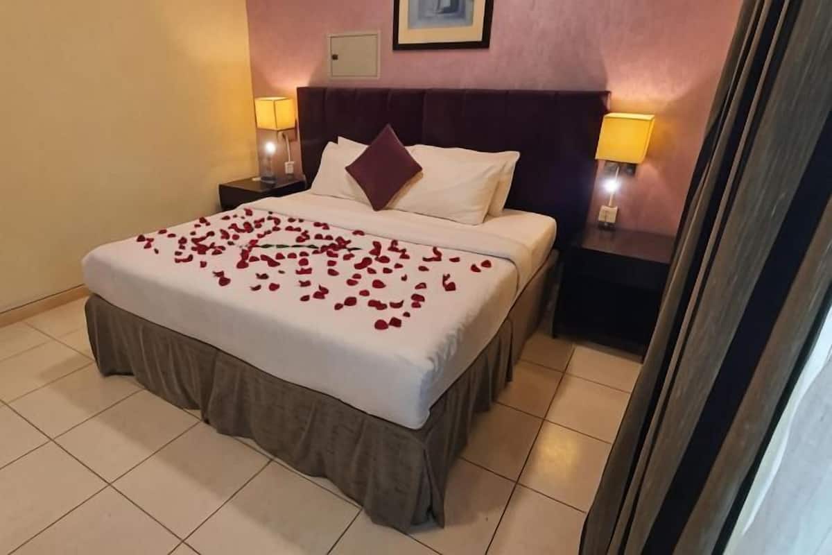 Cama bem arrumada com colcha branca e saia marrom, duas almofadas brancas, cabeceira escura e duas luminárias de cabeceira com abajures amarelos.