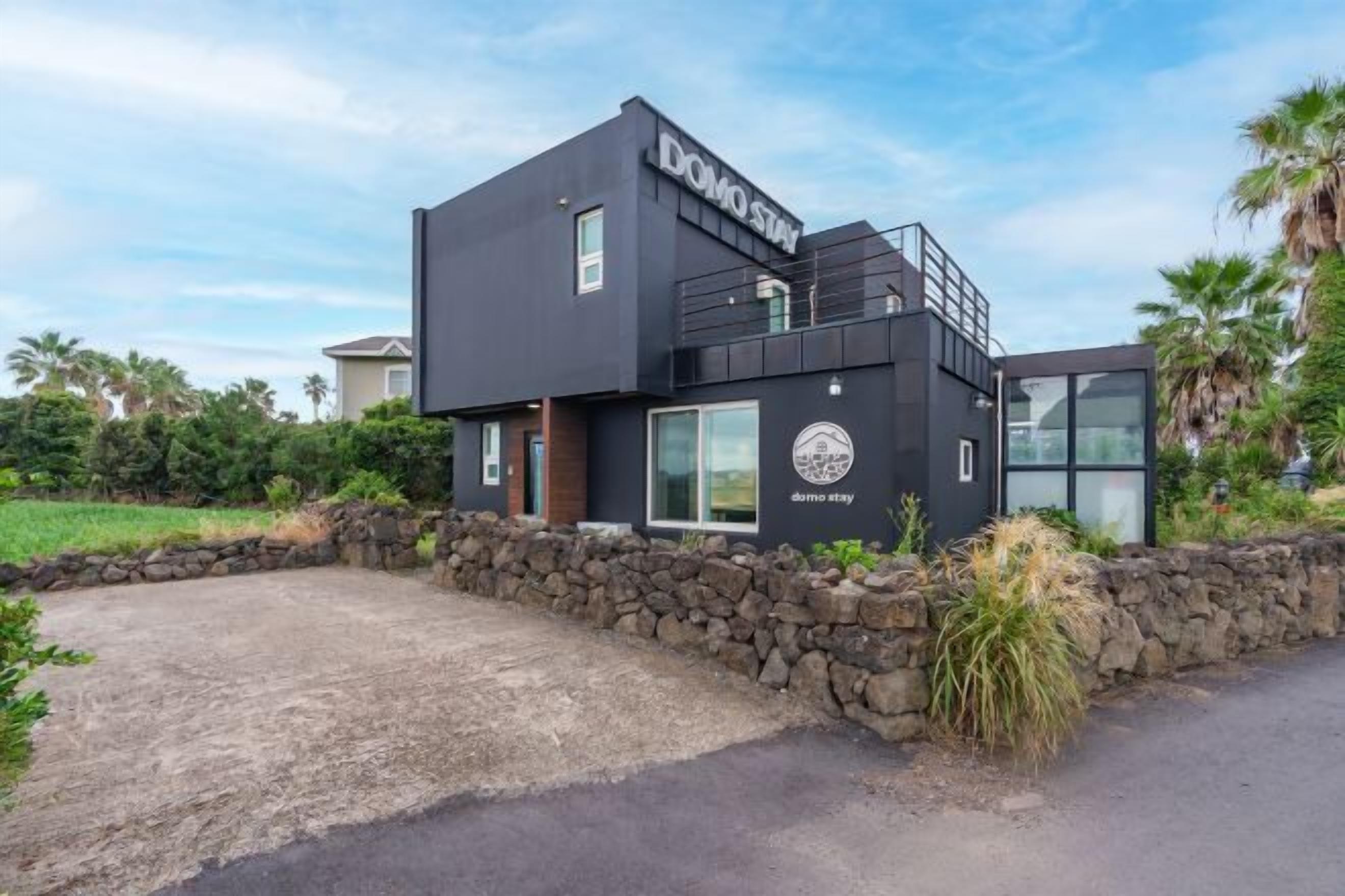 A modern black building with the name "DOMO STAY" on the facade, a stone wall in front, and a clear sky.