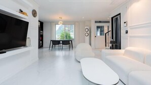 Ein modernes Wohnzimmer mit einem Flachbildfernseher, weißen Sofas, einem Essbereich mit Tisch und Stühlen und einem großen Fenster mit Blick auf Grünflächen.