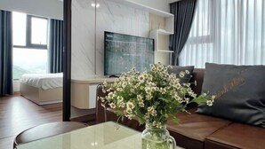 Ein modernes Hotelzimmer mit einem Glastisch, einer Couch mit Zierkissen, einem Flachbildfernseher, einem Bett und einem großen Fenster mit Vorhängen.