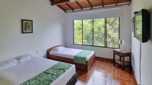 Habitación con dos camas, techo de madera, ventana con vista a follaje y una pequeña mesa.