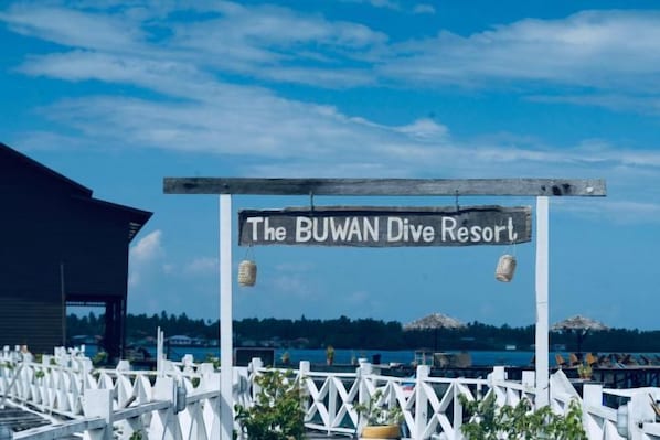 Señal para el Buwan Dive Resort, con una valla blanca y vista al mar.