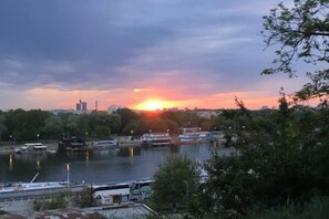 Um pôr do sol sobre um rio, com barcos, uma ponte e edifícios ao fundo.