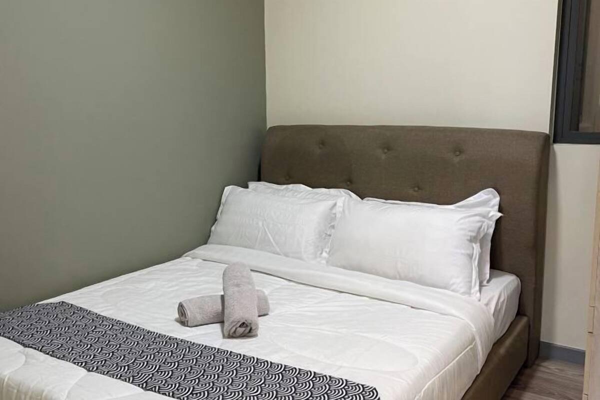 Ein ordentlich bezogenes Bett mit weißen Leinen und einer gemusterten Tagesdecke, ein braunes Polsterkopfteil und ein graues Handtuch auf dem Bett.