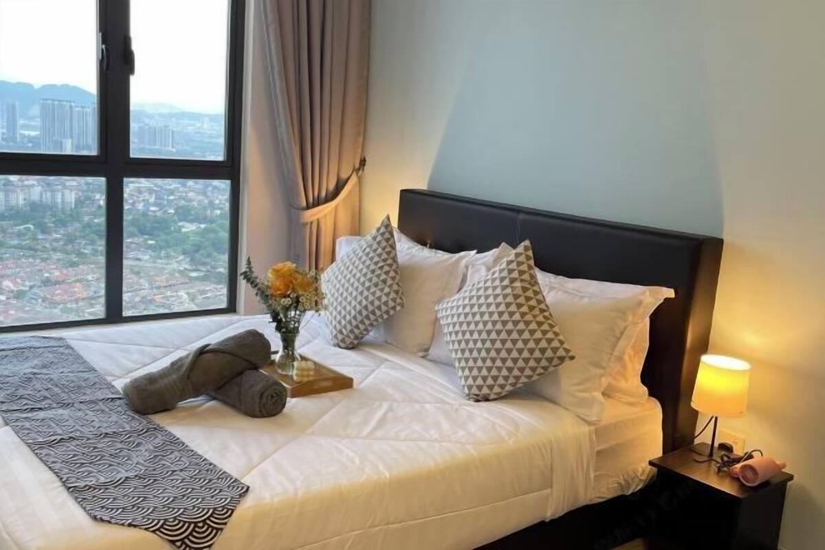 A neatly made bed with patterned pillows, a bedside lamp, and a view of a cityscape through a large window.