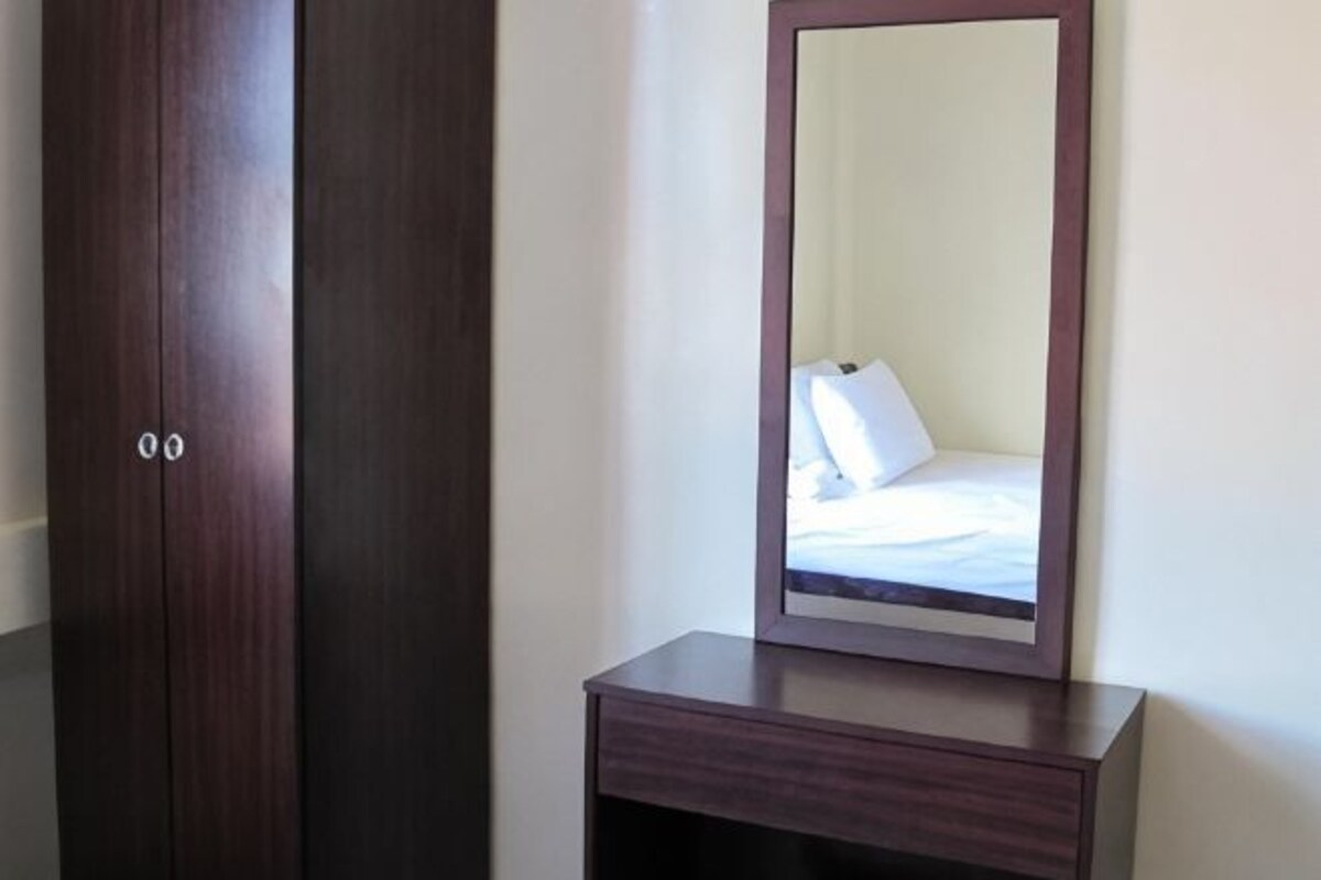 A dark wooden wardrobe with a mirror above it, reflecting a bed with white bedding.