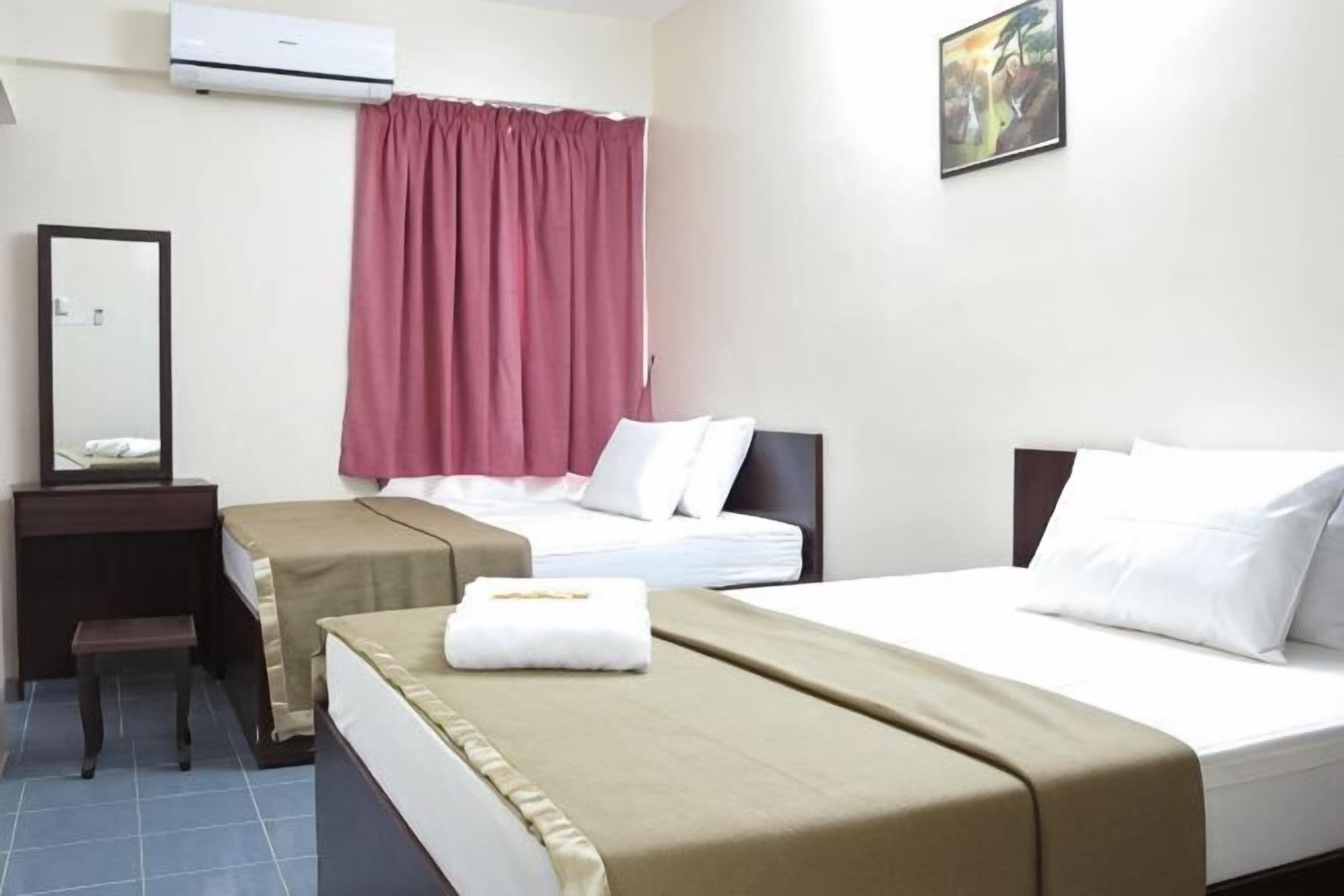 A hotel room with two beds, a desk, a chair, a mirror, a window with curtains, and an air conditioner.