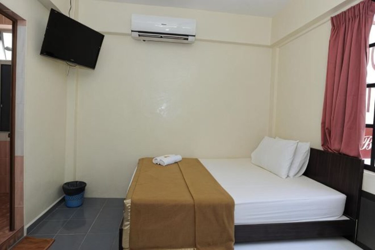 A single bed with a brown cover, a white pillow, and a white blanket. There is a black television mounted on the wall, an air conditioning unit, and a window with a red curtain.