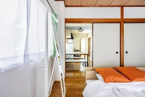 A bedroom with a bed, an orange pillow, a wooden floor, a white wardrobe, and a dining area with a table and chairs. - Nishikameari 2 chome (Tokyo)