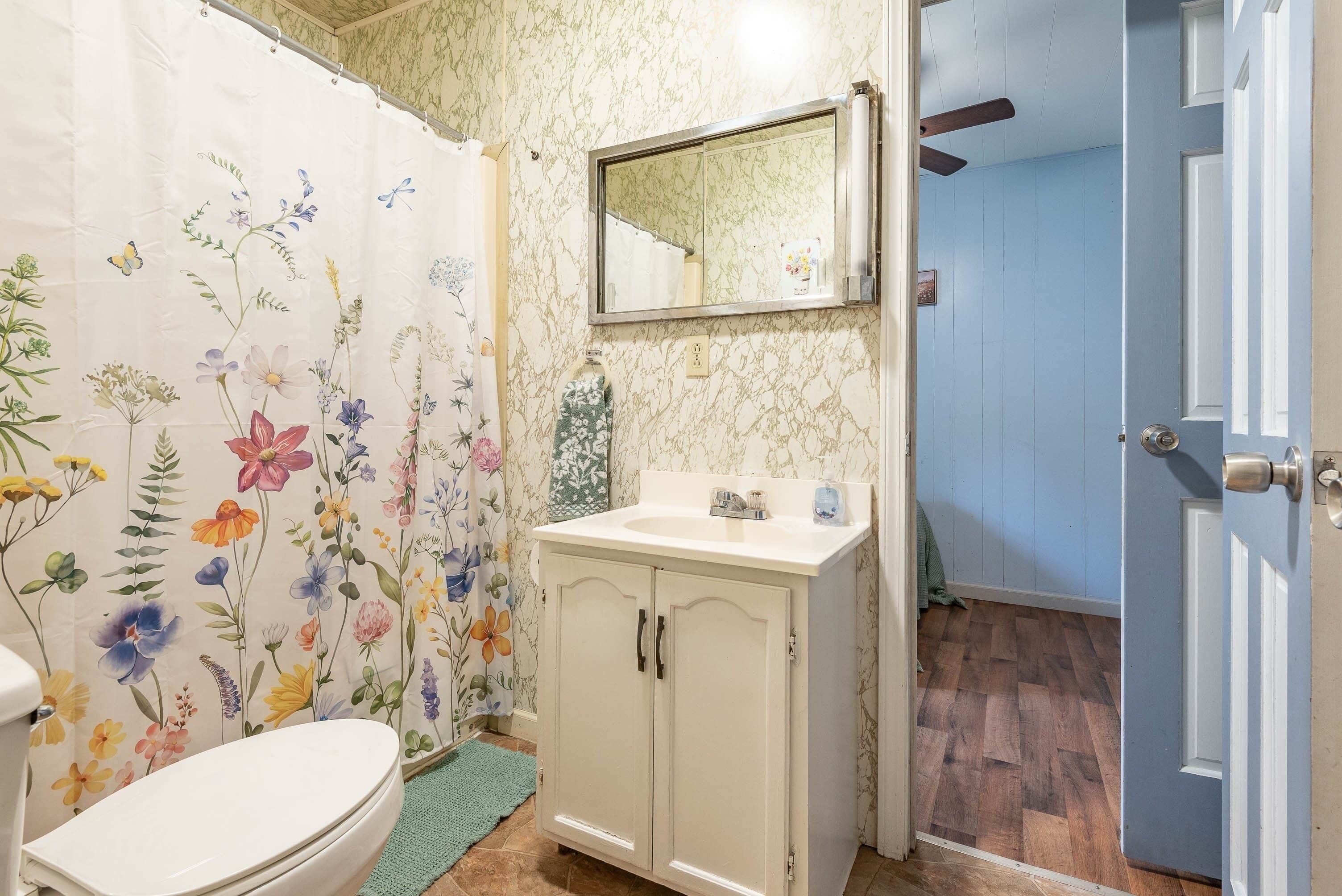A bathroom with a floral shower curtain, a toilet, a sink with a mirror, and wooden flooring.