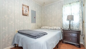 Une chambre à coucher avec un lit, une table de chevet avec une lampe, un papier peint à motifs floraux et un panneau décoratif portant l’inscription « Foi, Espoir et Amour ».
