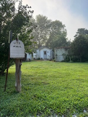 Une boîte aux lettres du service postal américain se trouve dans une cour verdoyante, avec un petit bâtiment blanc en arrière-plan.