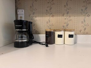 Un comptoir de cuisine avec une machine à café, une prise de courant et trois récipients étiquetés pour le café, la crème et le sucre.