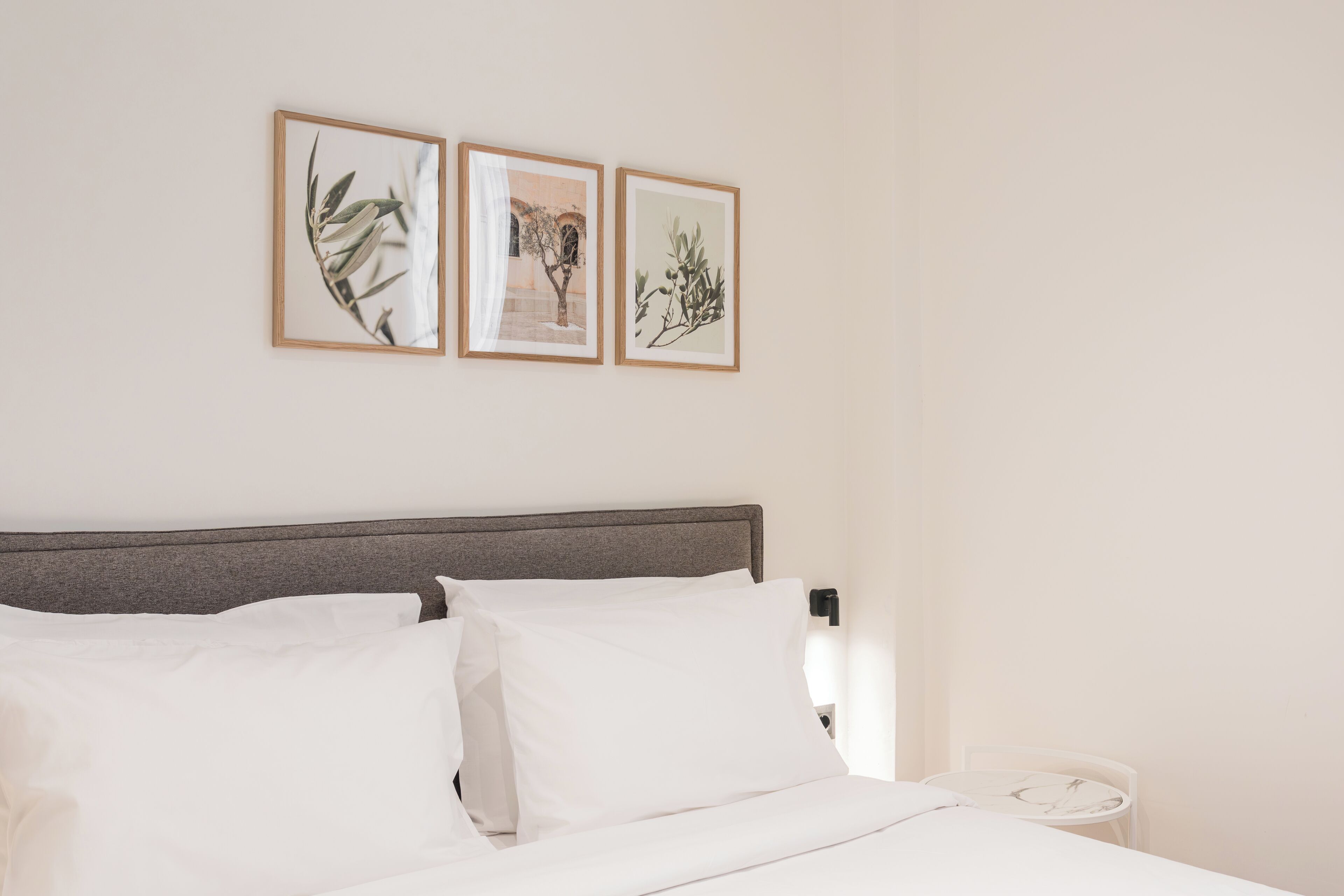 A neatly made bed with white linens, a wooden headboard, and a nightstand with a lamp. There are three framed botanical prints on the wall above the bed.