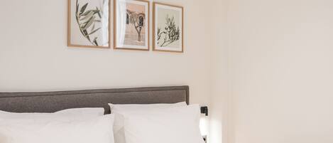 A neatly made bed with white linens, a wooden headboard, and a nightstand with a lamp. There are three framed botanical prints on the wall above the bed.