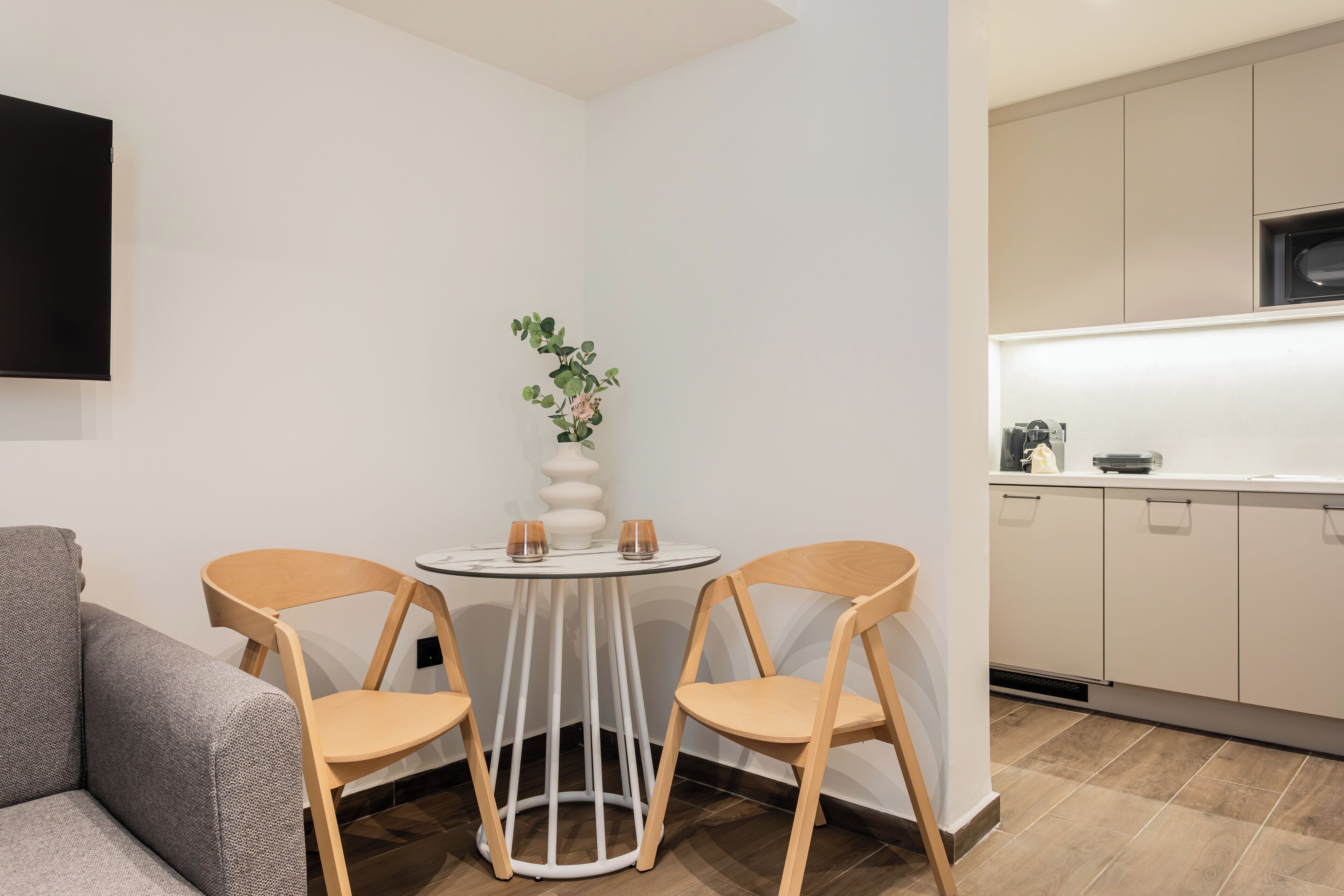 Een ruimtelijk ingerichte zithoek met een bank, een klein rond tafeltje met twee stoelen, een potplant en een keukenhoek met kasten en een magnetron.
