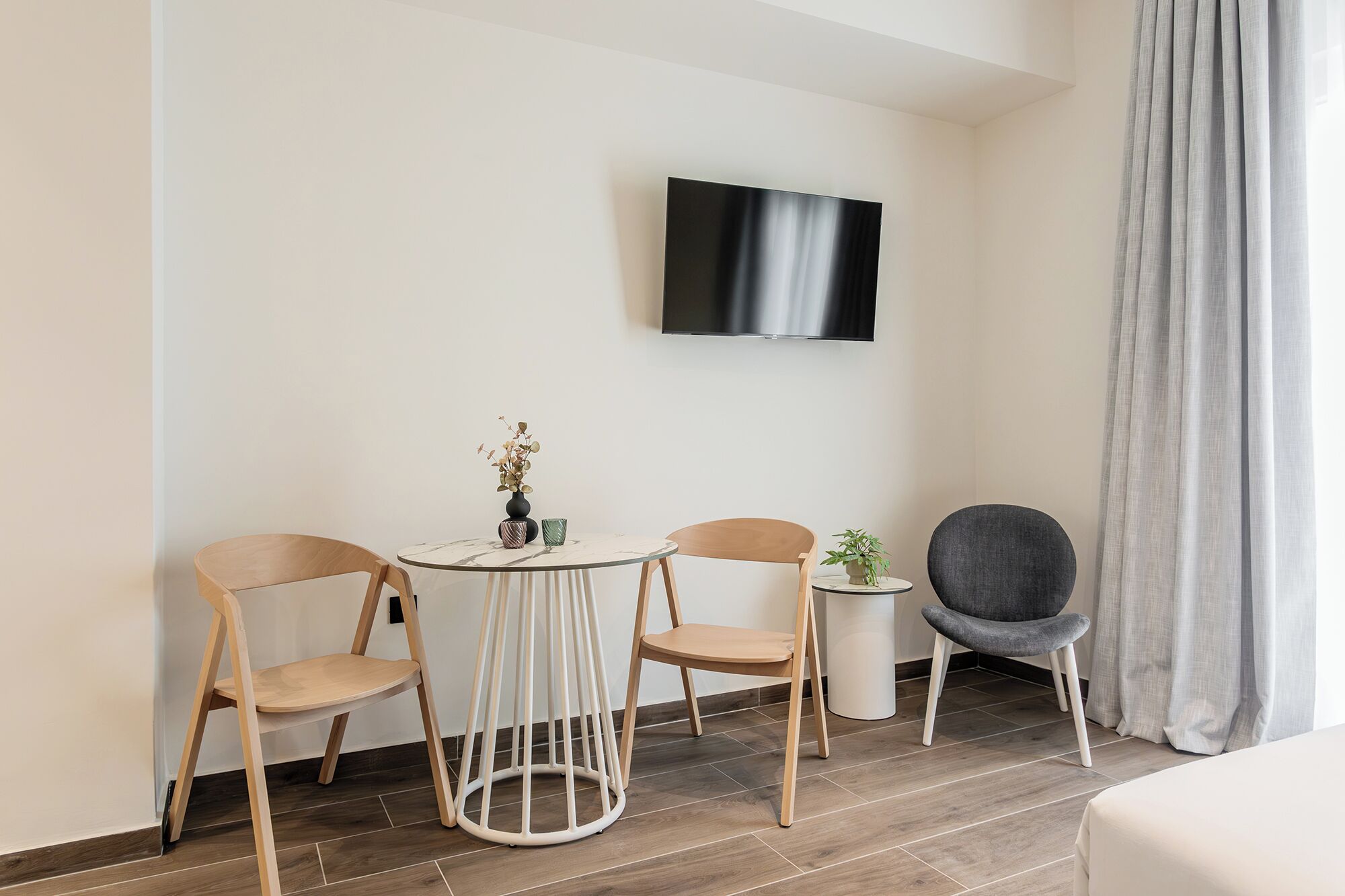 A modern room with a round table, two chairs, a potted plant, a grey armchair, and a flat-screen TV mounted on the wall.