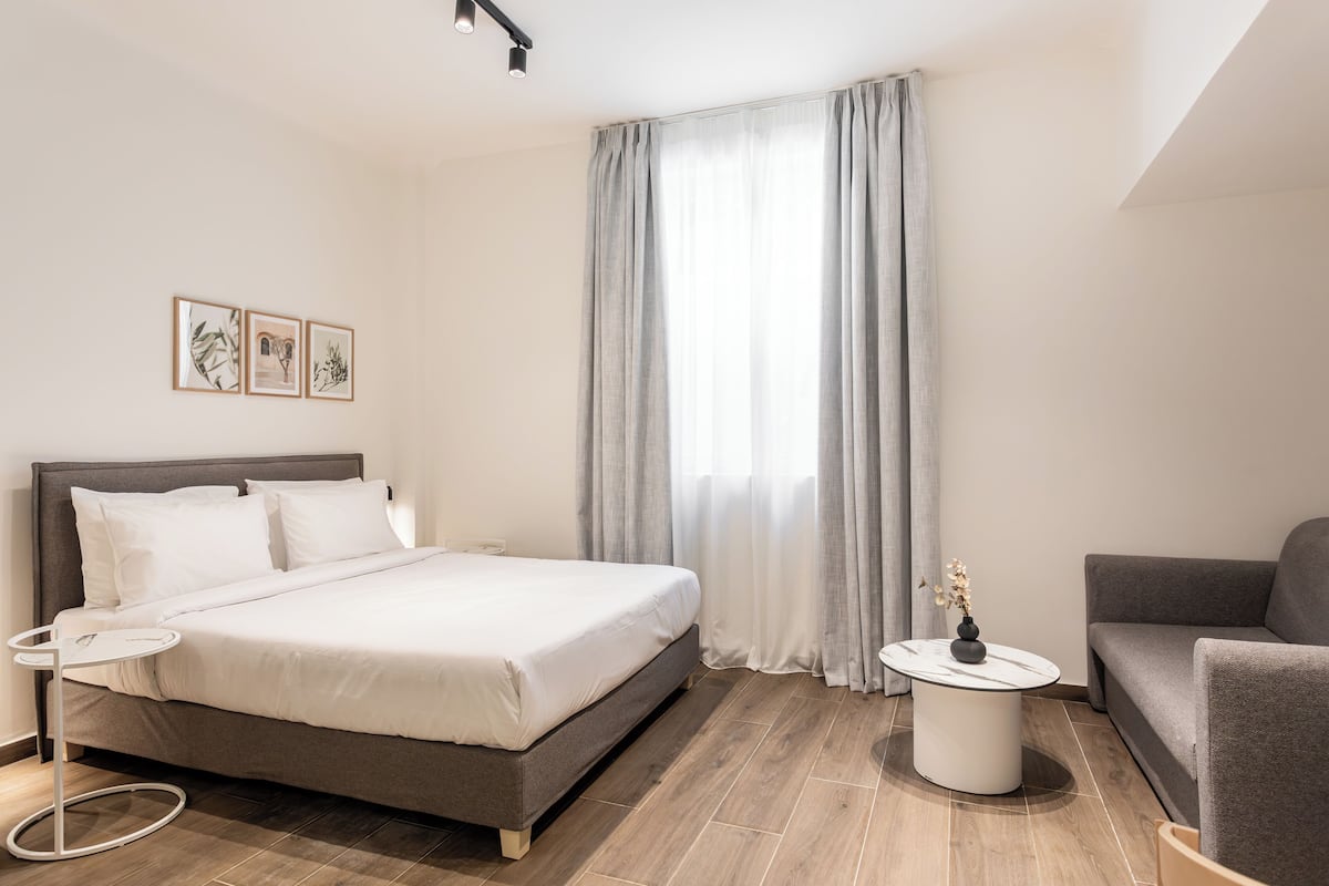 Ein modernes Hotelzimmer mit einem großen Bett, einer Couch, einem kleinen runden Tisch und einem Fenster mit Vorhängen.