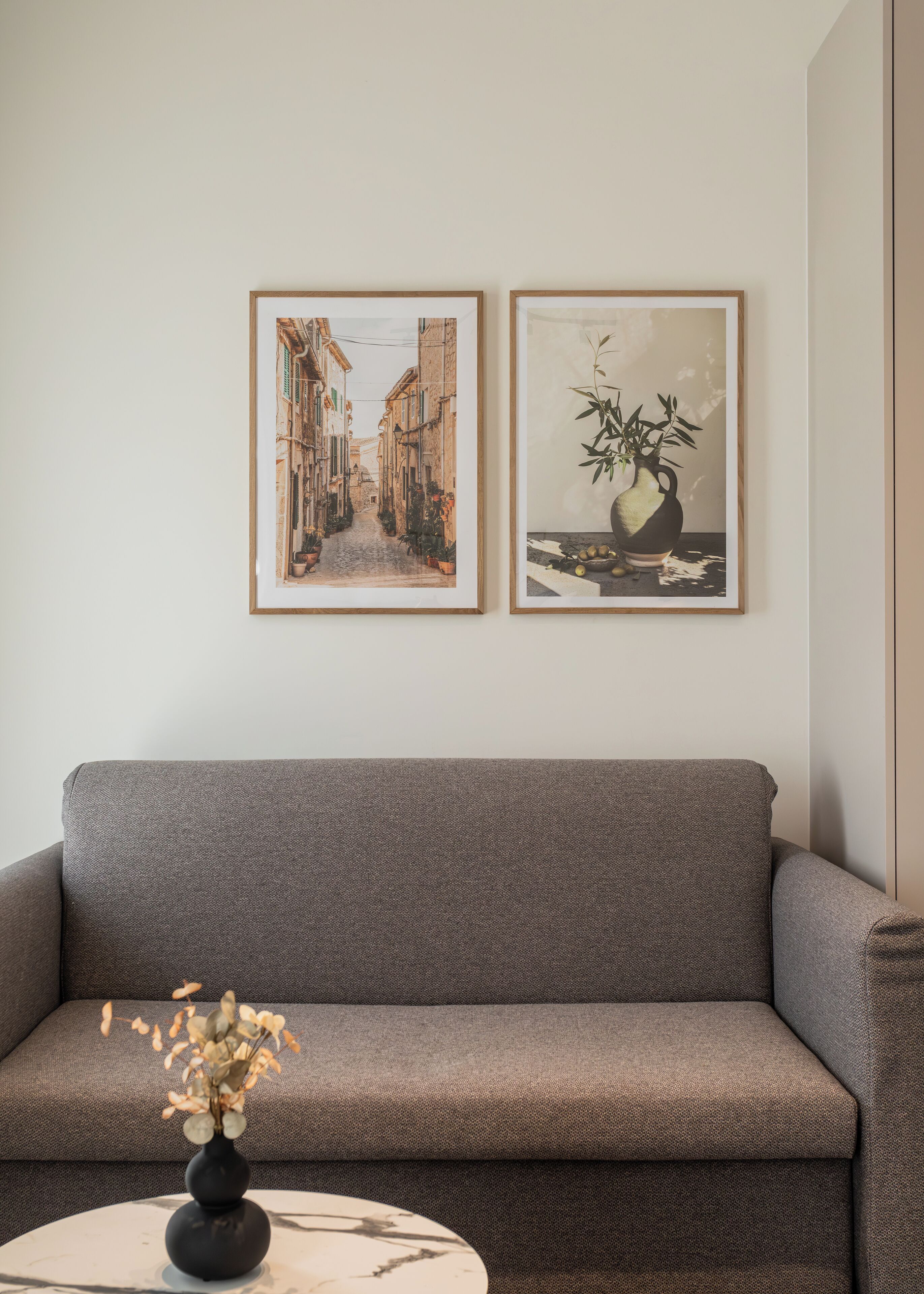A living room with a grey sofa, a small round table with a vase, and two framed pictures on the wall.