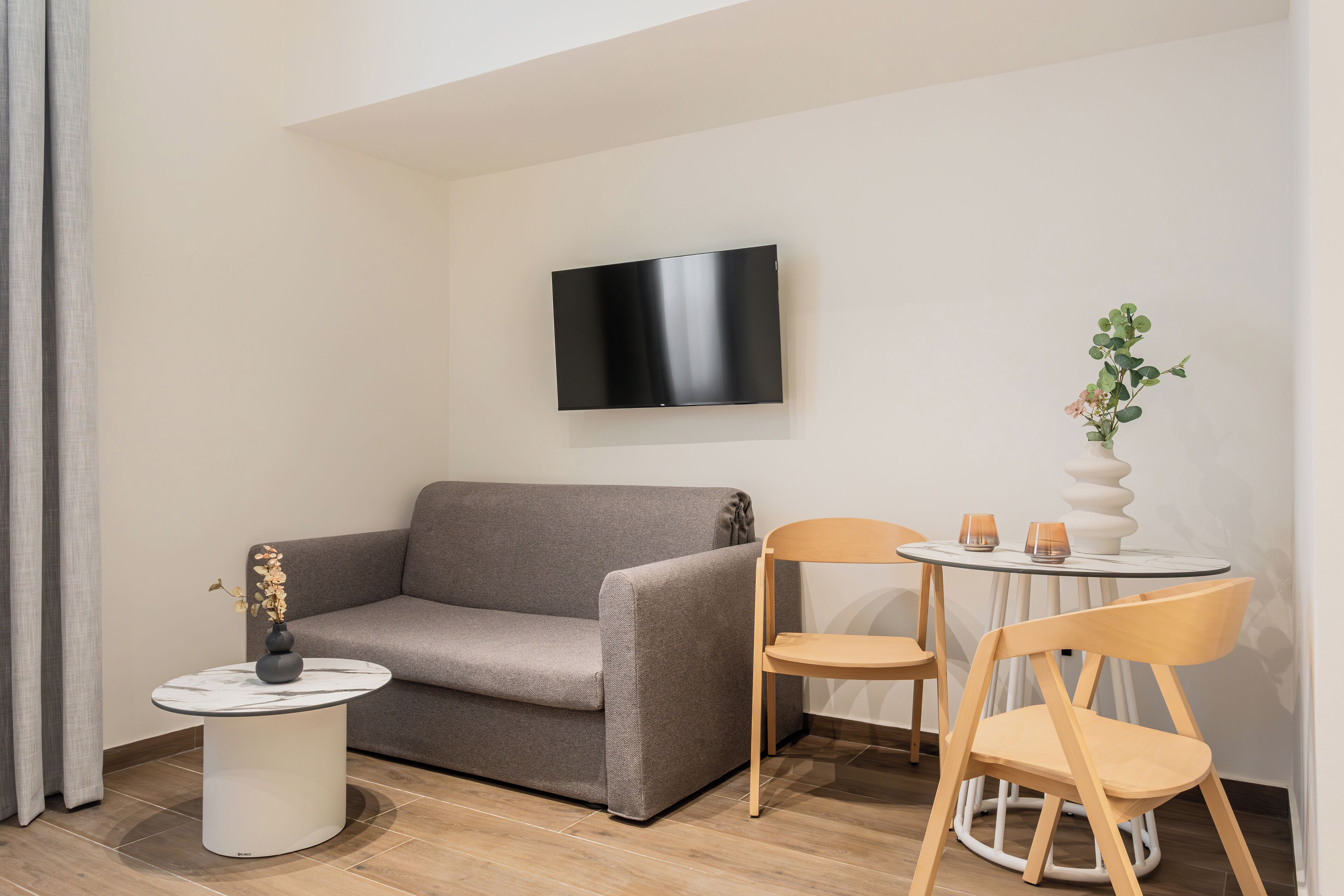 A modern living room with a sofa, a small round table, two chairs, a flat-screen TV, and a vase with flowers.