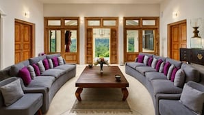 A spacious living room with a large wooden coffee table, two grey sofas with purple cushions, and wooden doors leading to a garden. - The Divine Villa (Heenatigala)