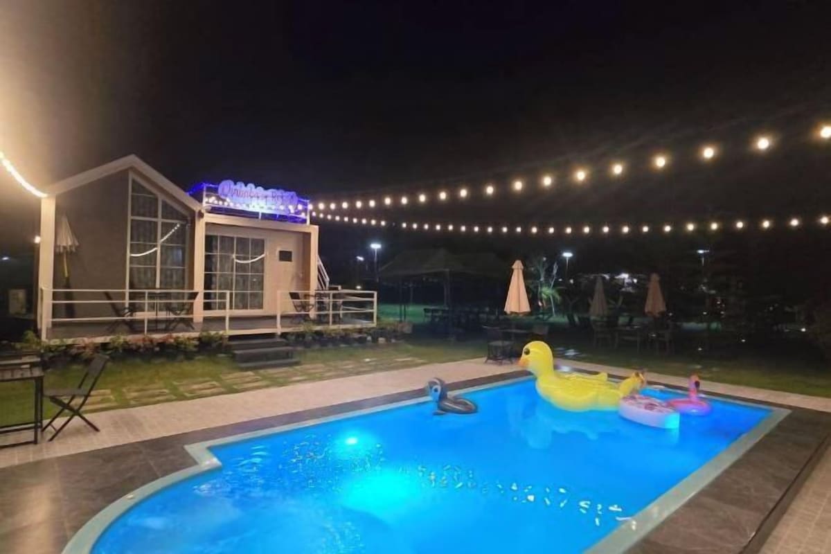 A swimming pool with inflatable toys, a building with a neon sign, and string lights in the background.