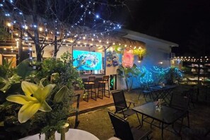 Un espace extérieur aménagé pour les repas, avec des tables et des chaises, un coin bar doté d’un téléviseur et des guirlandes lumineuses.