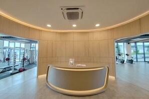 A reception area with a curved desk, a fire alarm, and a view of the gym equipment. - K Avenue by Butter House @Sunset Seaview Cozy (Donggongon)