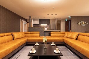 A modern living room with a sectional sofa, a coffee table with cups, and a kitchen area in the background. - THE SKY (Sapporo)