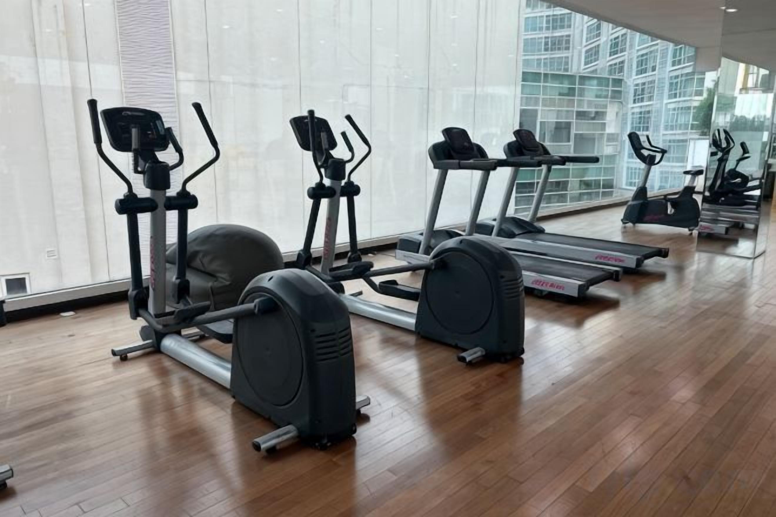 A gym with cardio machines, including treadmills and elliptical trainers, on a wooden floor with large windows.