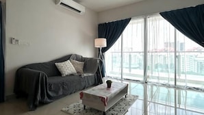 A living room with a sofa, a coffee table, a lamp, and a view of buildings through large windows. - M9 Prime Suite@H20 Medalla CittaMall SubangAirport (Petaling Jaya)