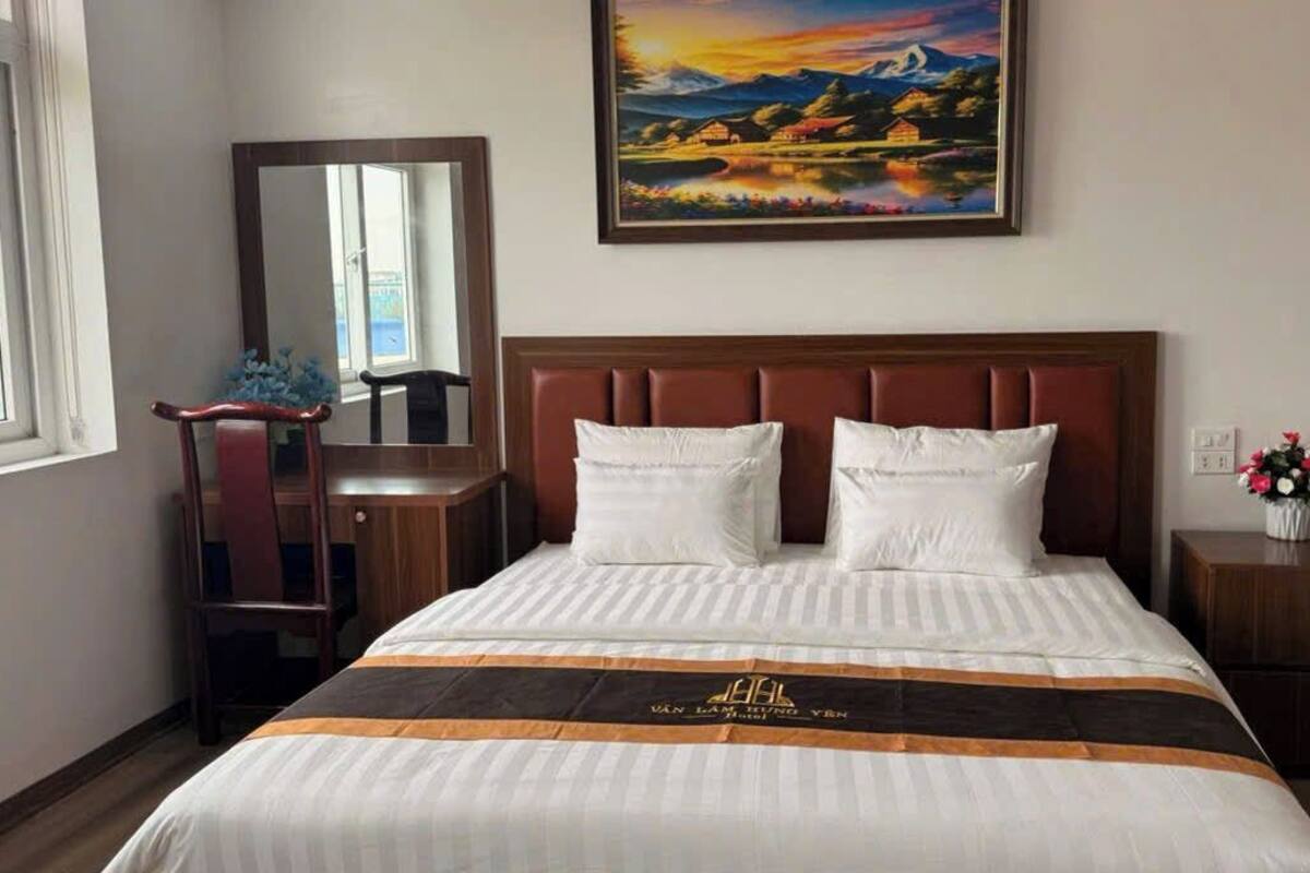 A hotel room with a bed, a nightstand, a mirror, a window, and a painting on the wall.