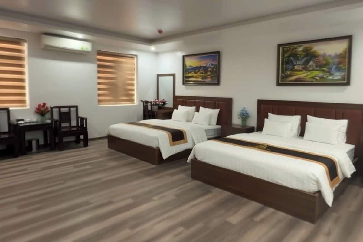 A hotel room with two beds, a desk, a chair, and two windows with blinds.