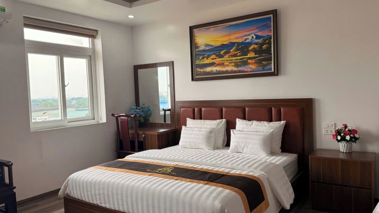A hotel room with a large bed, a nightstand, a mirror, a window with a view, and a painting on the wall.