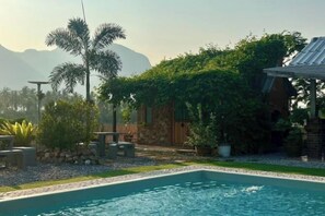 Une piscine bordée de pierres, entourée d’un jardin où poussent différentes espèces de plantes et délimitée par une clôture en bois.