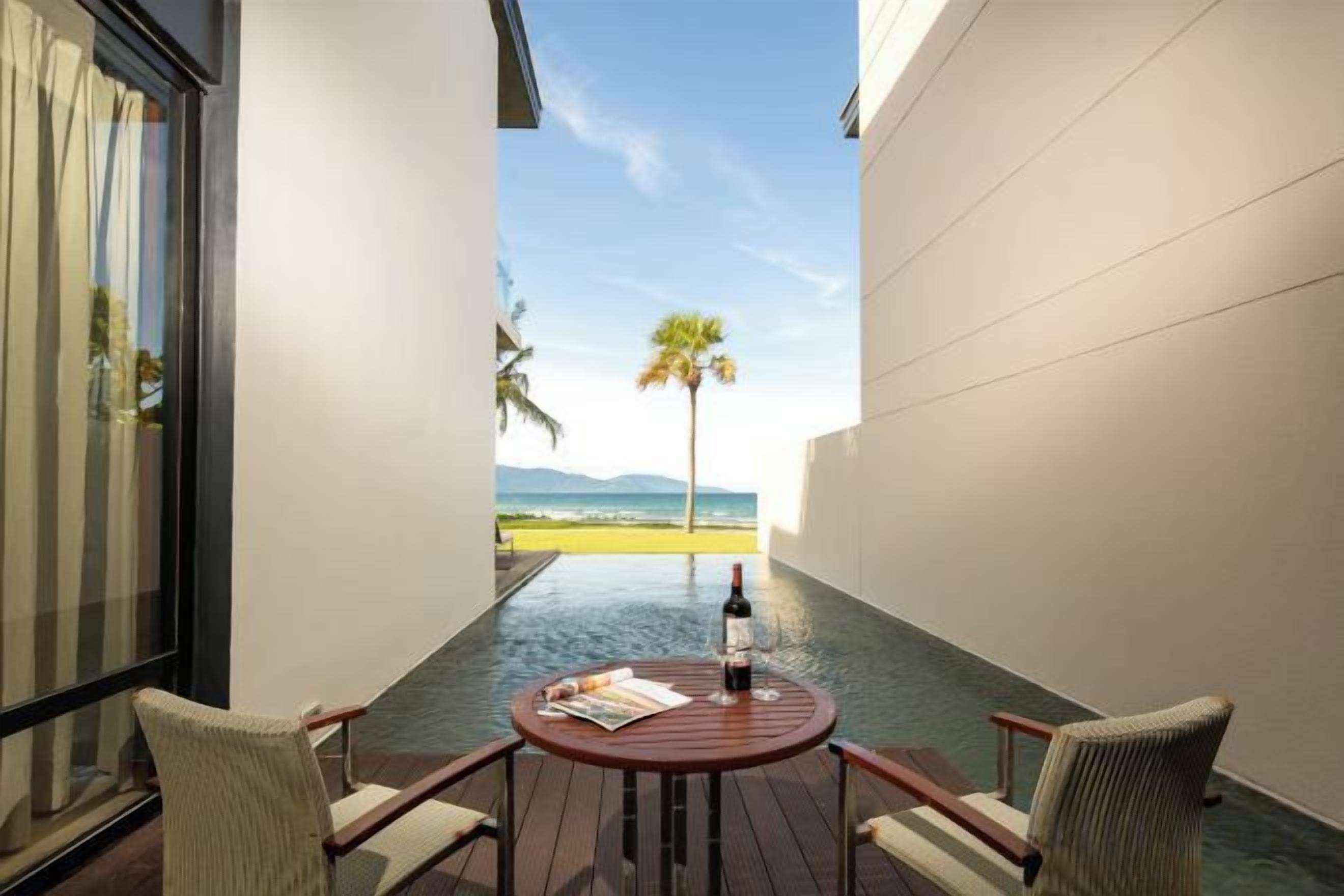 A balcony with a table, chairs, and a bottle of wine, overlooking a palm tree and a distant view.