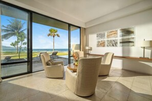 A spacious room with a large sliding glass door leading to a balcony with a view of the ocean and palm trees. There are several chairs and a small table with a vase of flowers. - Beachfront Villas Resort Danang (Da Nang)