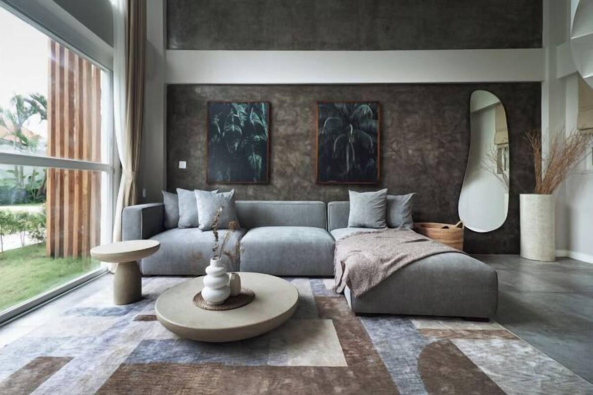 A modern living room with a grey sofa, two wall-mounted paintings, a round coffee table, and a large window with a view of greenery.