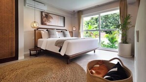 A modern bedroom with a large bed, a small bedside table, a hanging light fixture, a large window with a view of greenery, and a potted plant. - A.K.A Tropicana Bingin Uluwatu (Bali)